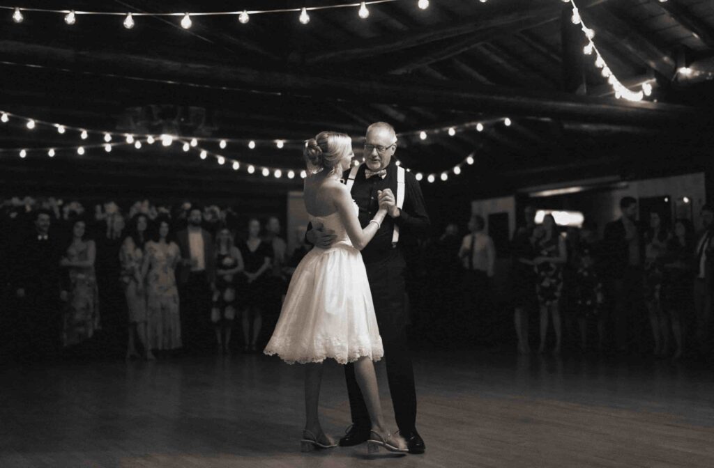 First dance of the Bride in a short white wedding dress and father of Bride with guests watching in the background