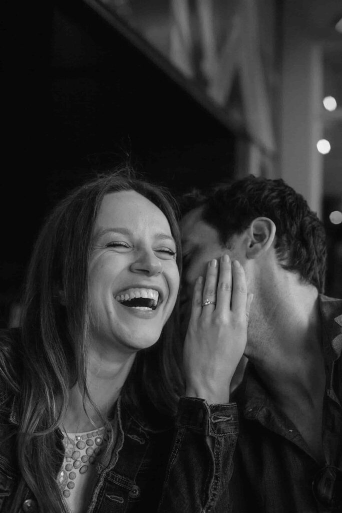 A man is whispering in a woman's ear while she holds his face and laughs while showing off her engagement ring