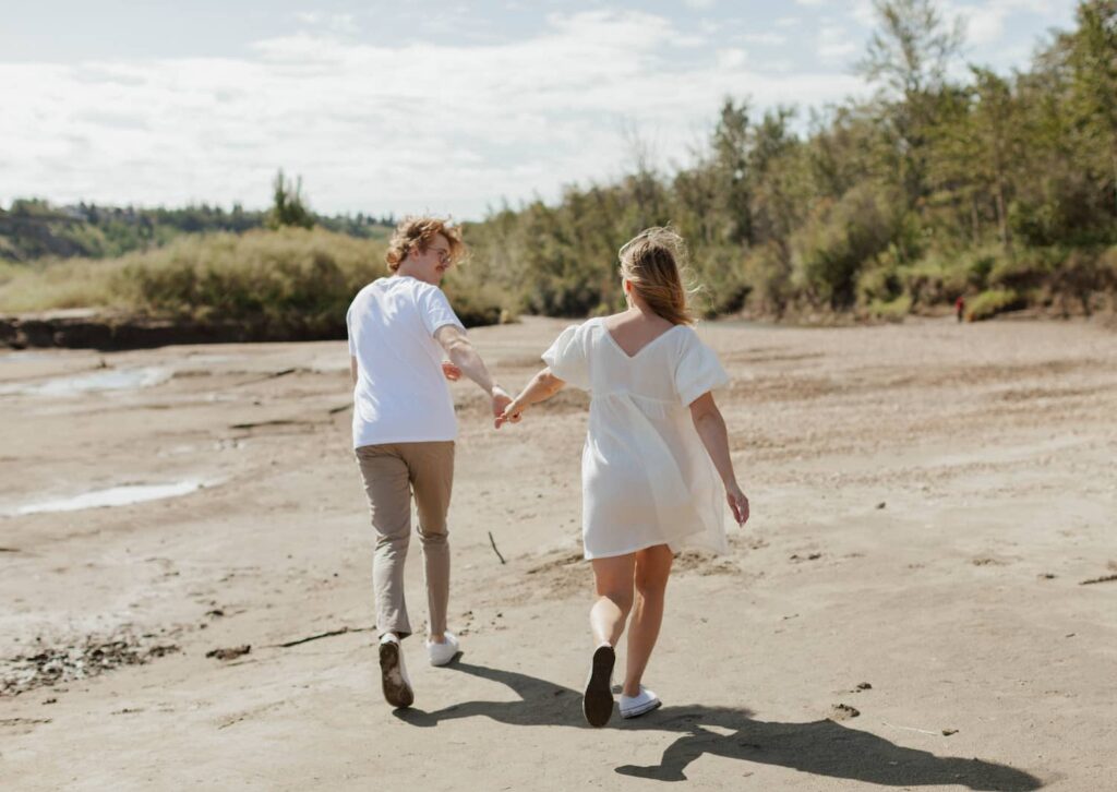 A couple running across a sandy beach holding hands and looking at eachother. They are engaged