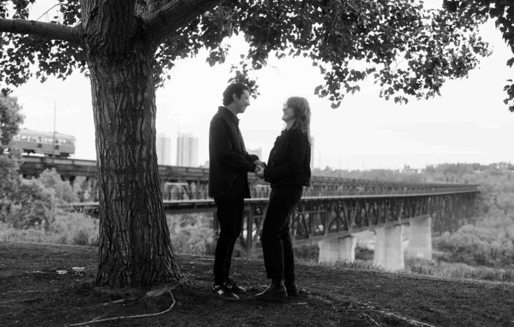 Couple standing under a tree holding hands and looking at each other with the High Level Bridge in the background