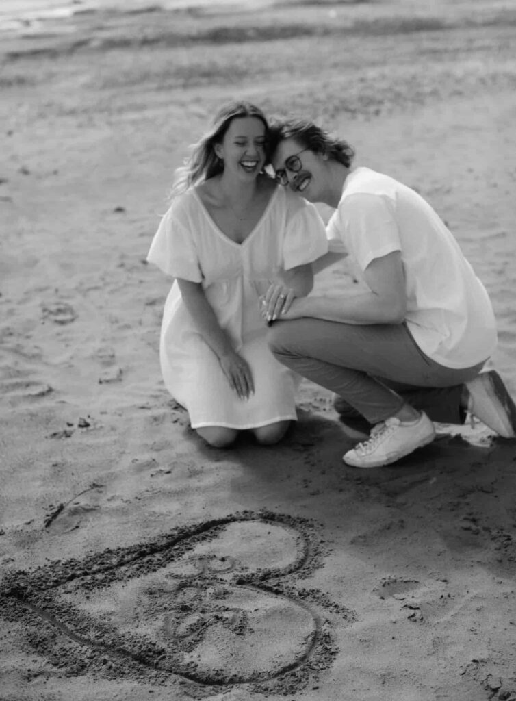 An engaged couple together who are laughing on a sandy beach in Edmonton, Alberta with a heart drawn in sand