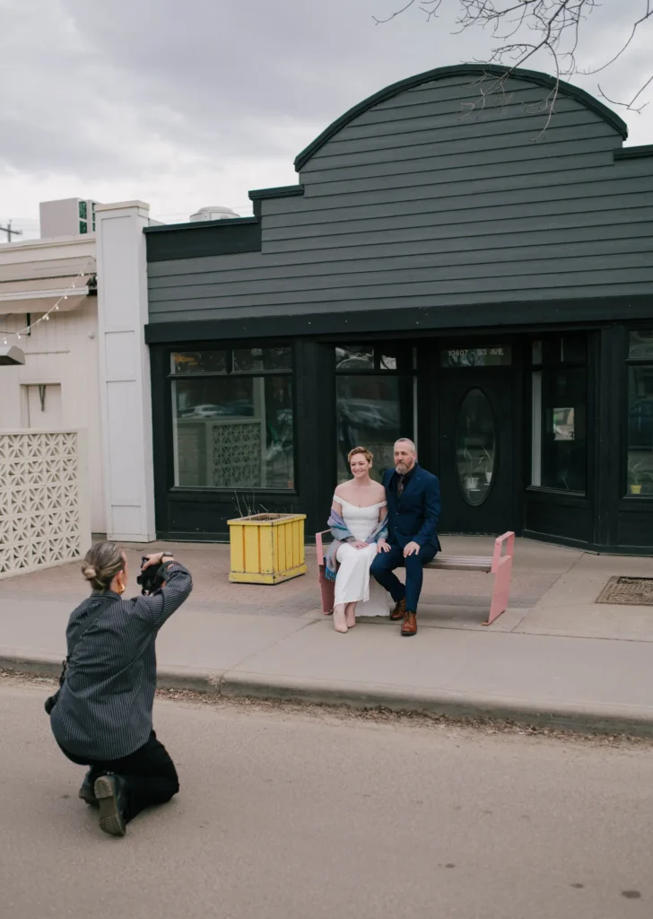 woman photographing bride and groom on a bence in front of a building