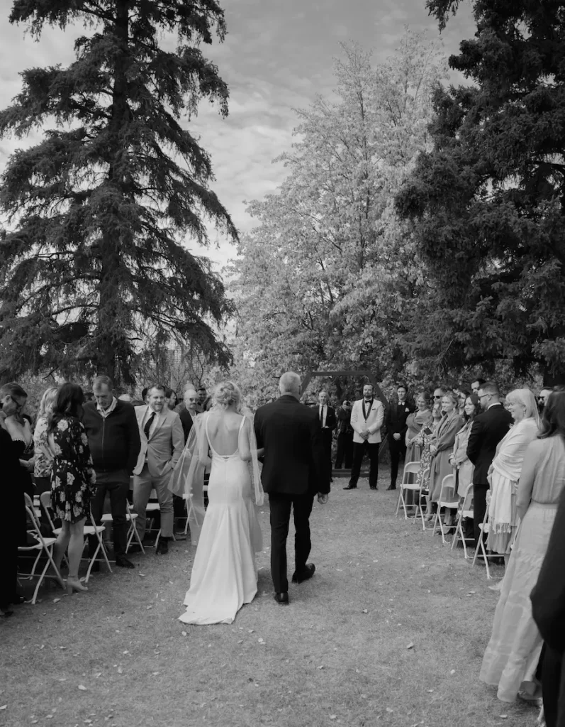 outdoor wedding ceremony where the brides father is walking her down the aisle towards the groom. There are people on either side sitting in chairs looking at the bride as she walks down the grassy aisle