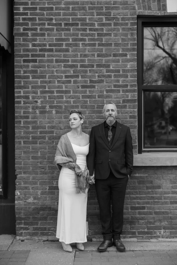 bride and groom standing against a brick wall while bride looks out and groom looks at camera