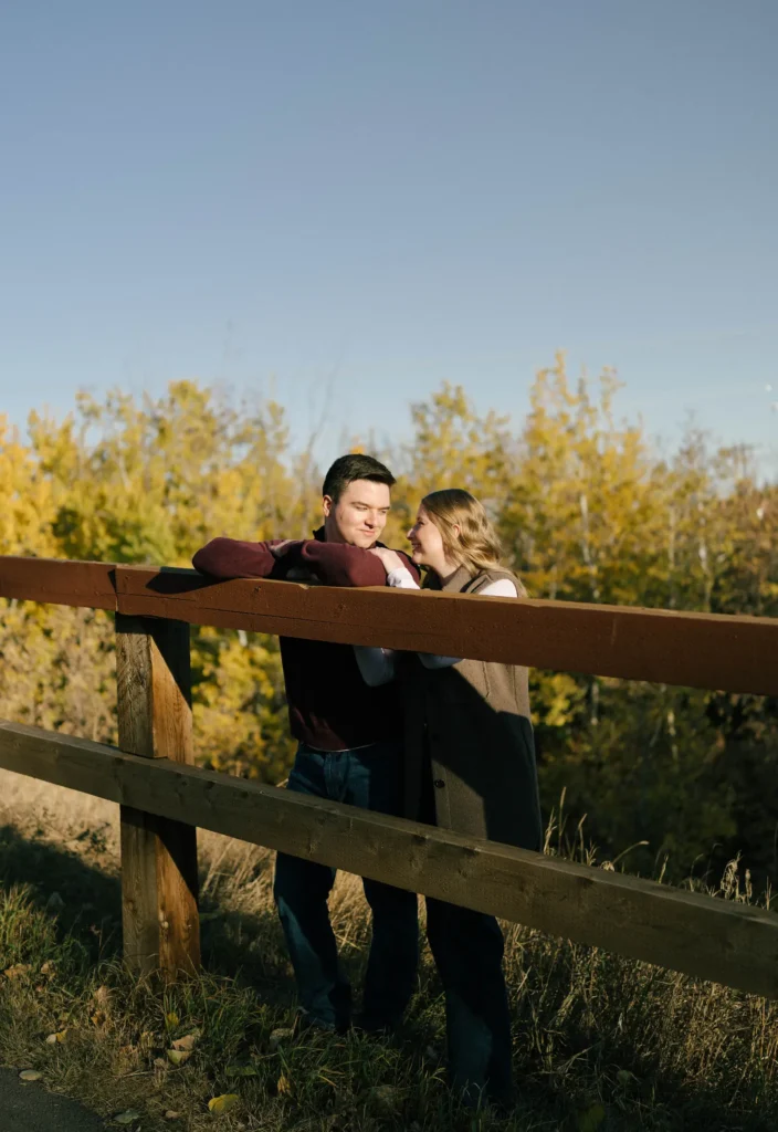 capilano park engagement photos of a couple standing against a fence looking at eachother with a blue sky in the background