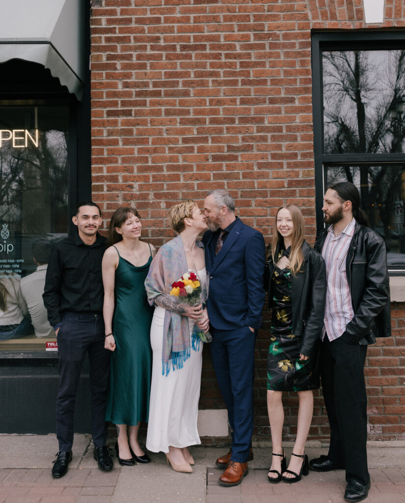 Five people stand in a row against a brick wall as a couple in the center share a kiss. The woman holds a bouquet while the others smile and look toward them.