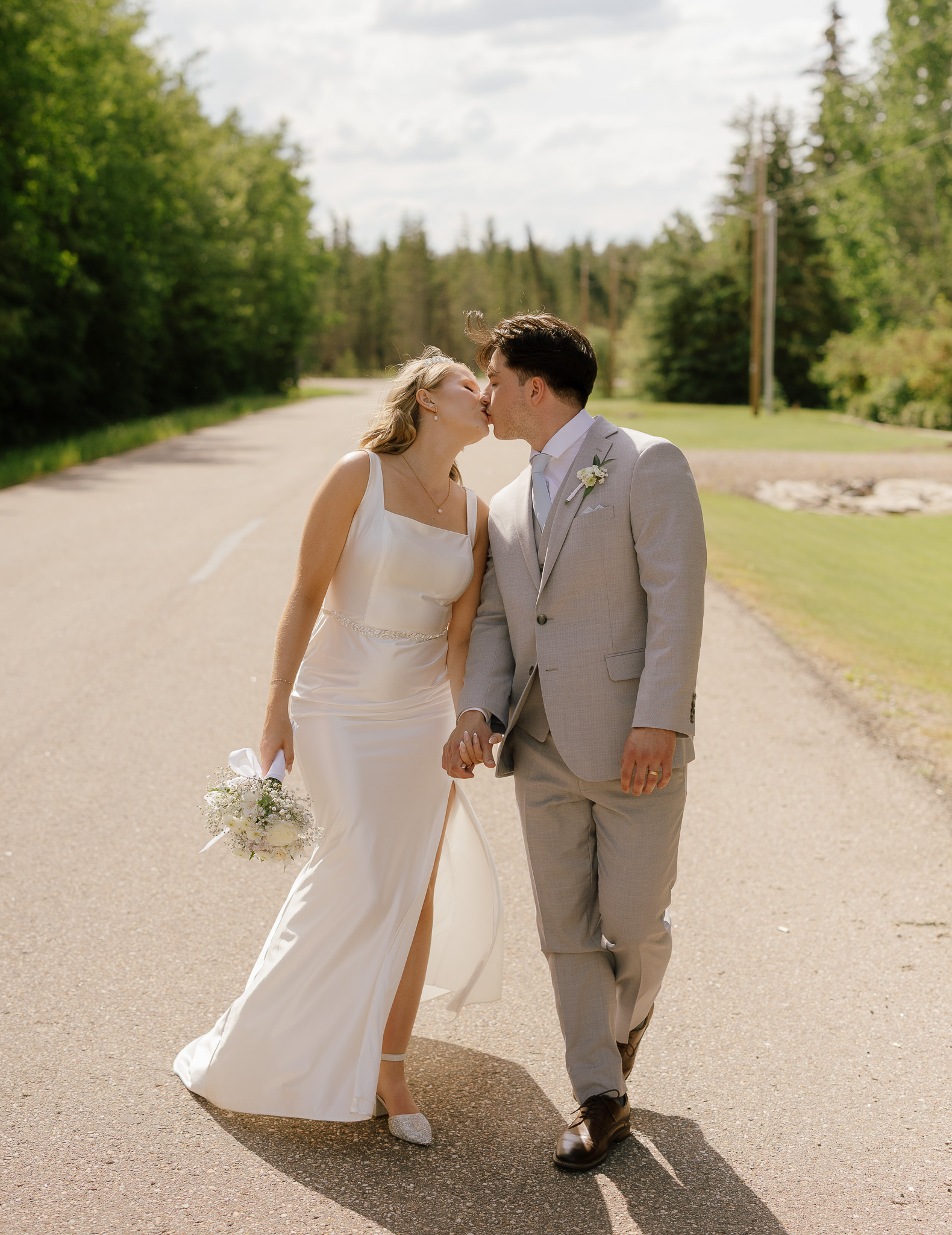 Bride and groom share a kiss while walking hand in hand down a quiet road during their Bonnyville, Alberta wedding.
