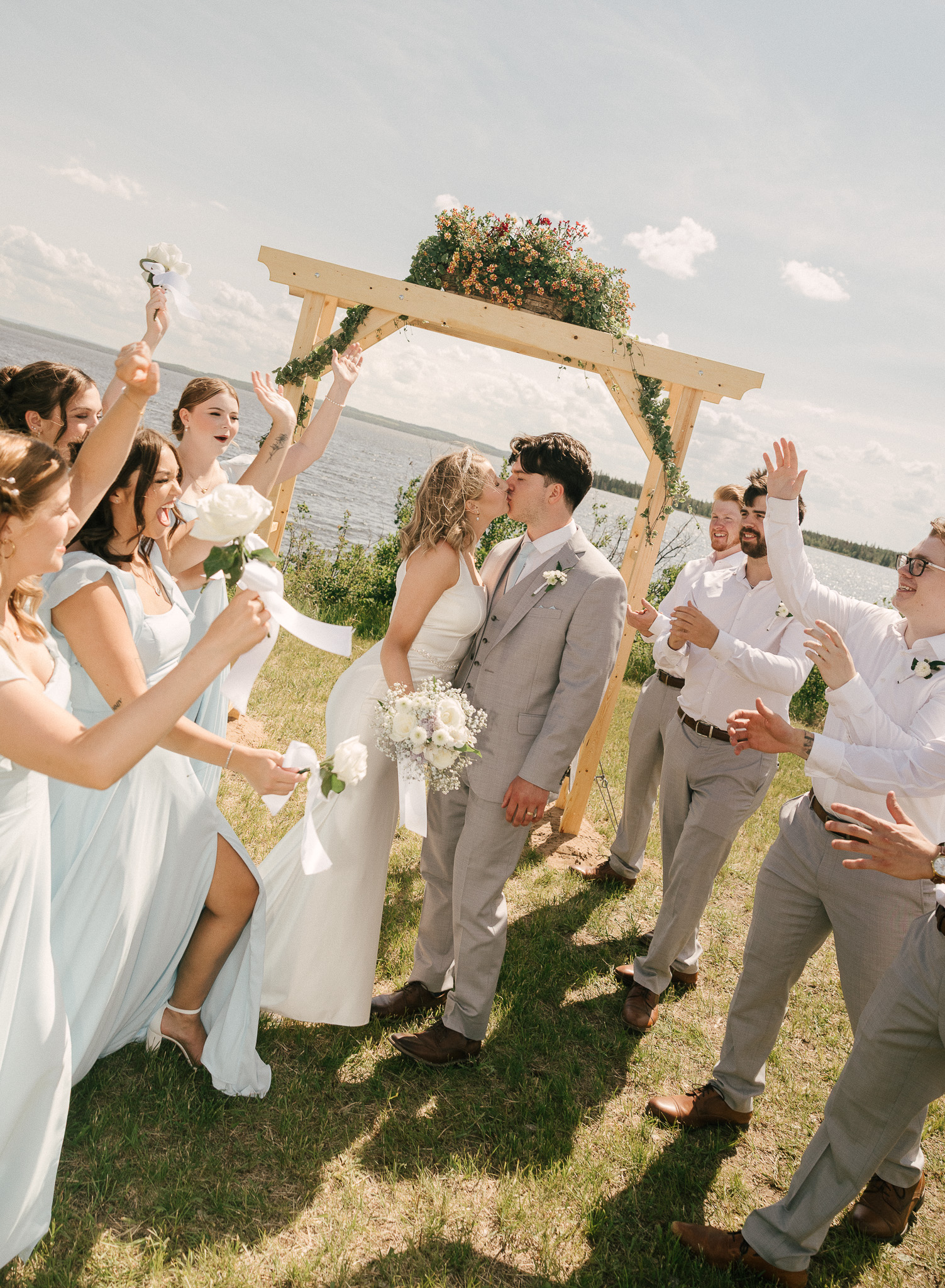 Bride and groom sharing a kiss surrounded by their cheering wedding party during an outdoor lakeside wedding ceremony near Bonnyville, Alberta.