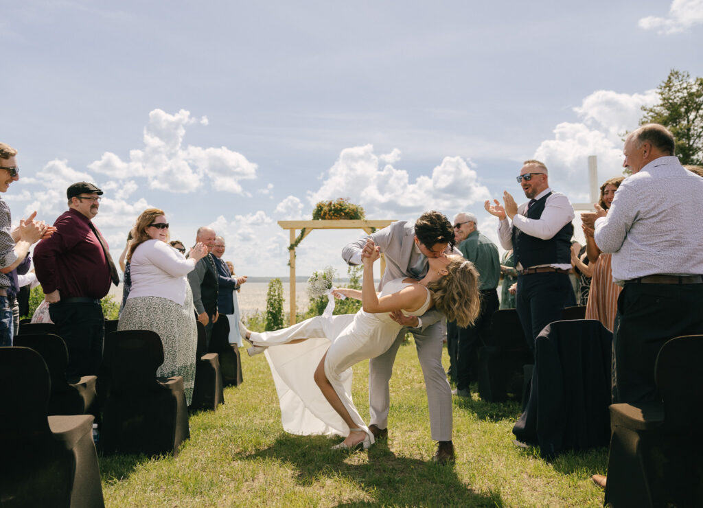 Newlyweds share a celebratory dip kiss while walking back down the aisle as guests applaud during an outdoor wedding ceremony in Bonnyville, Alberta.