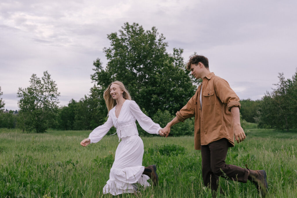 Engaged couple walking hand in hand through a grassy field during a summer engagement session at Terwillegar Dog Park in Edmonton, Alberta.
