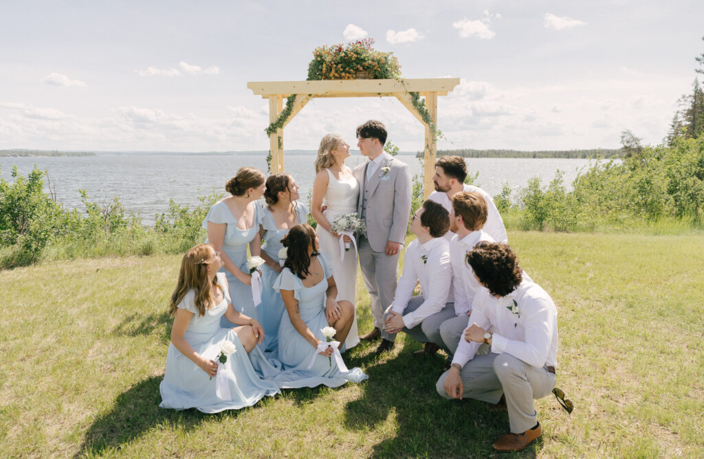 bridal party looking at bride and groom while they gaze into eachothers eyes with a large lake in the background