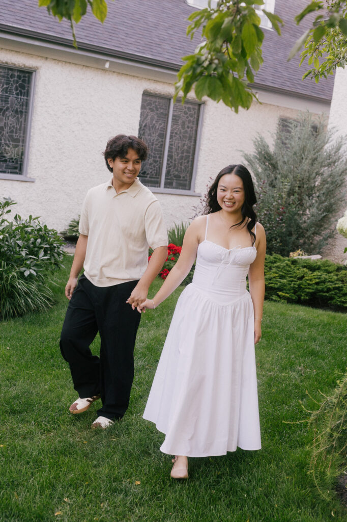 Engaged couple walking through a green garden next to a church in Wihkwentowin