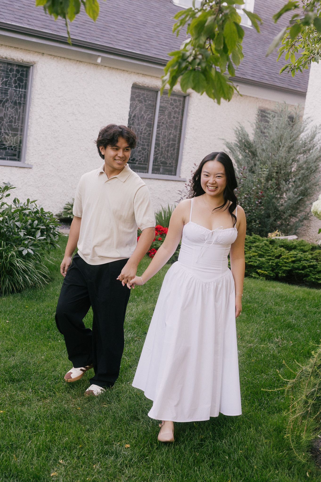 Engaged couple walking through a green garden next to a church in Wihkwentowin